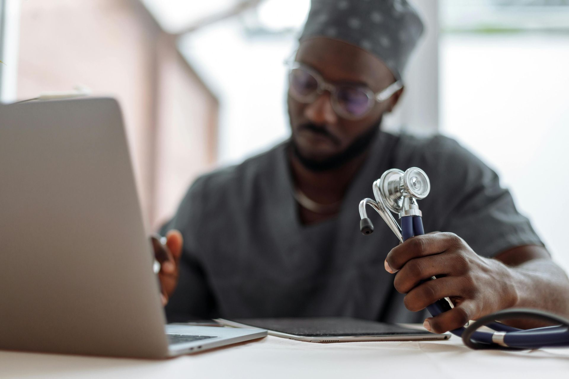 Focused African American doctor in scrubs working with laptop and stethoscope.